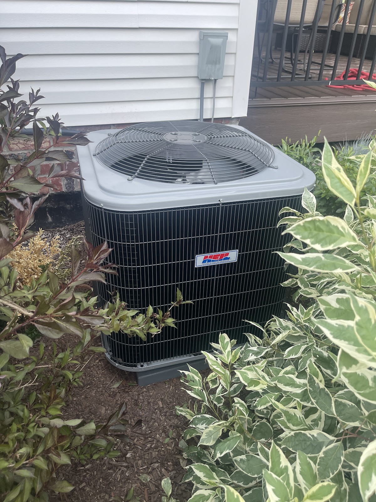 Residential HEI-brand central air conditioning condenser installed among landscaping beside a brick home